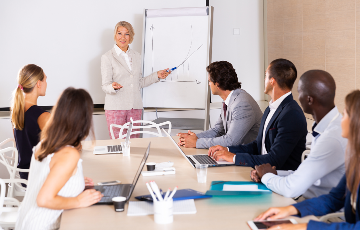 Woman giving a business presentation to an engaged group. She points out a hand-drawn graph.