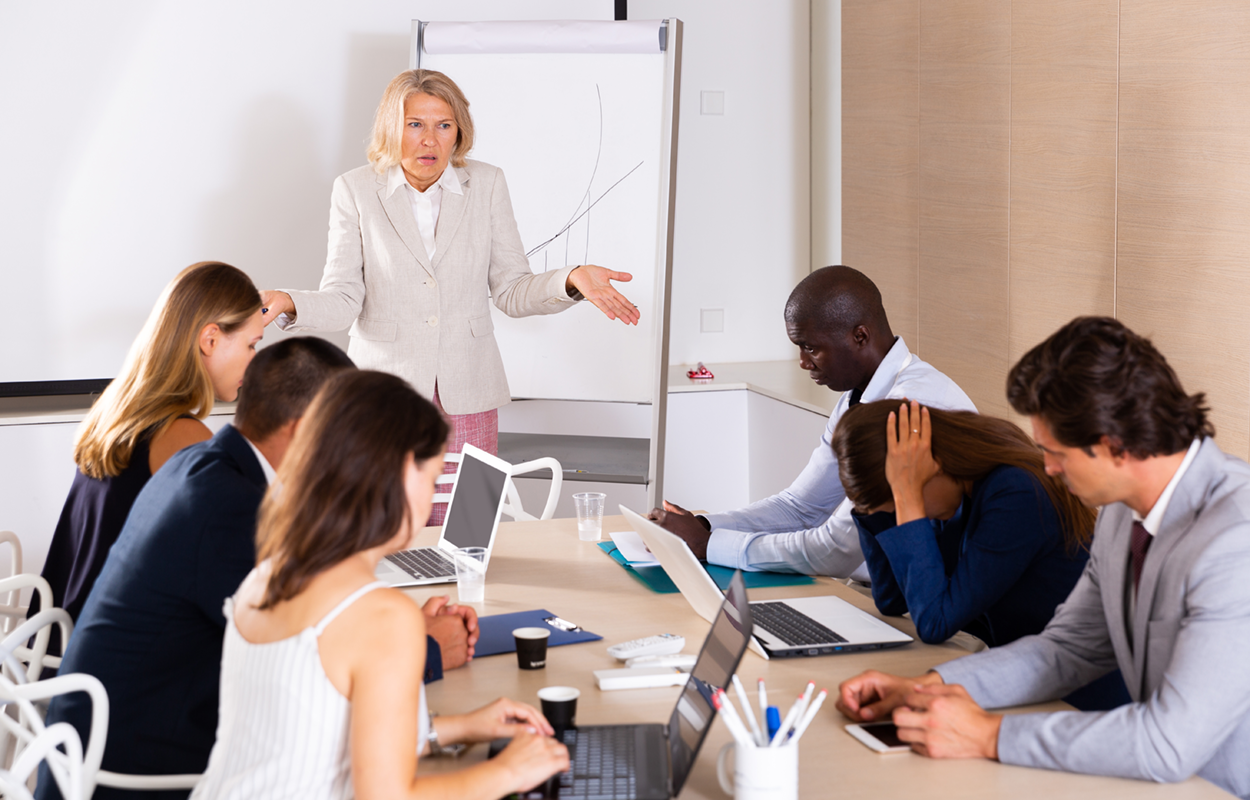 Woman giving a presentation to a group that seems disengaged or displeased by the presentation. She shrugs in frustration.