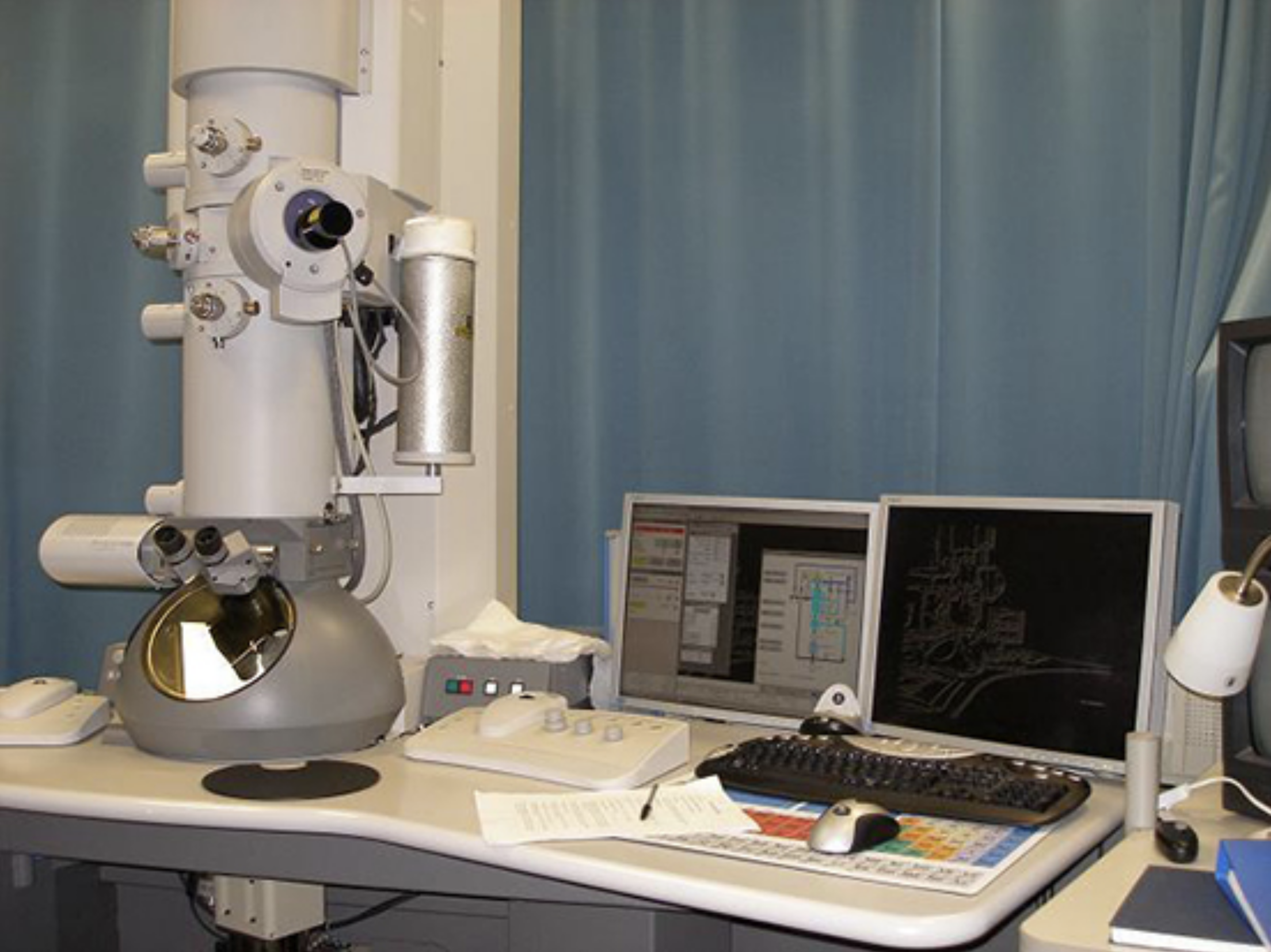 A photograph shows a transmission electron microscope. It is a large-tube shaped machine attached to a desk next to a computer.