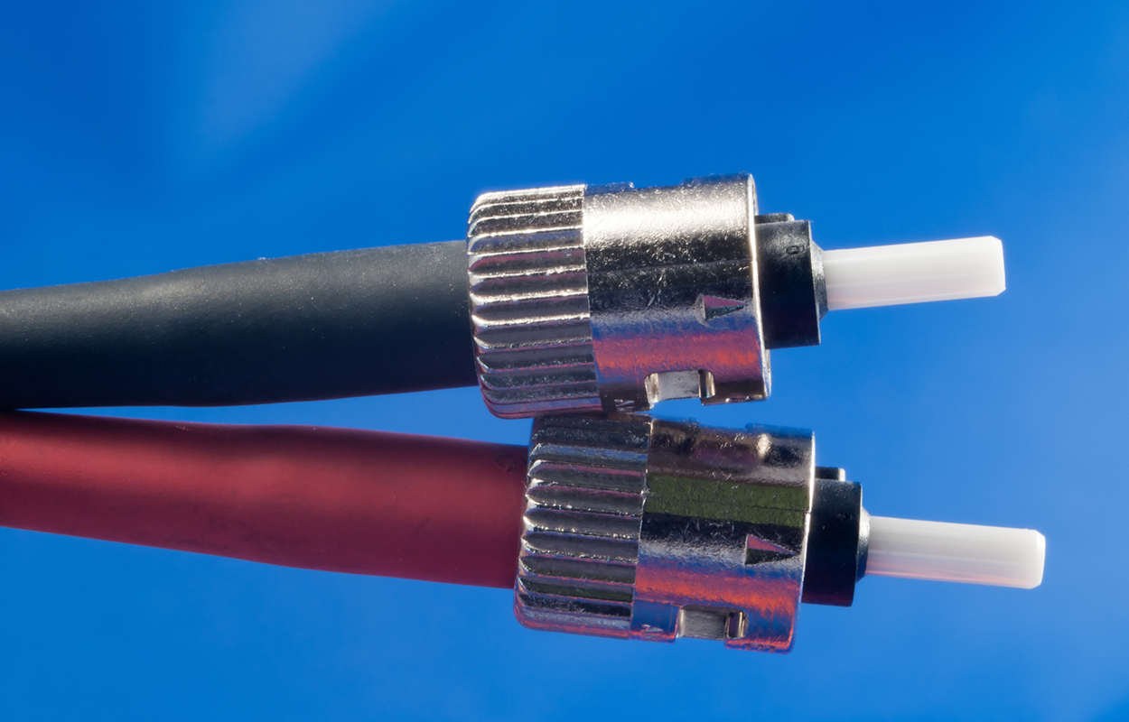 Two fiber-optic cables with S T connectors. Each connector has a metal screw-on part and a plastic tip. The cables are of different shades.