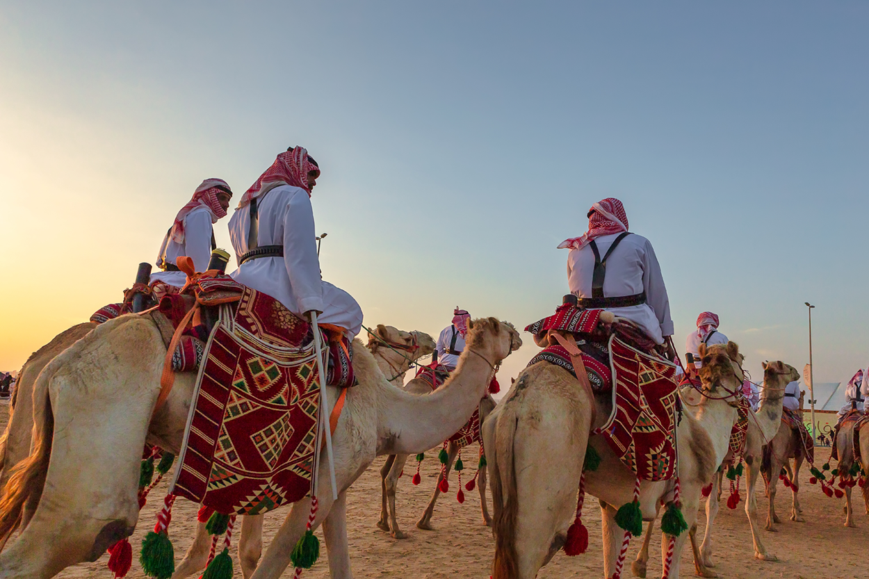 Individuals wearing all white clothing riding camels in a desert environment.