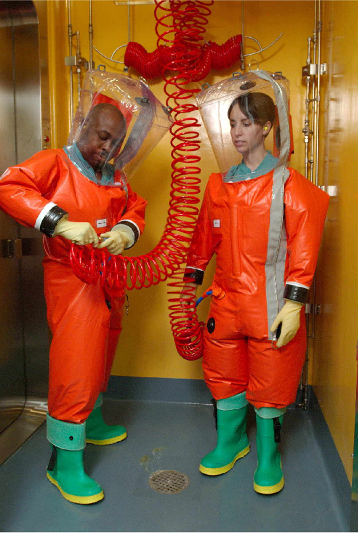 Two people are wearing bright orange suits with clear head covers and bright green boots with yellow soles. All parts of the suits are connected with no gaps. One person is attaching a long, bright orange, coiled air tube to the side of his suit and the other person has a similar tube already attached. the tubes extend upward to attach to something above the photo.