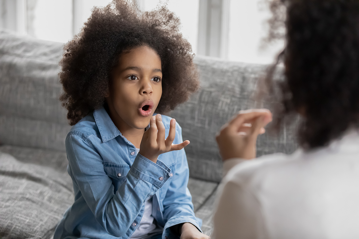A child sitting on a couch is articulating the “o” sound with their lips open in a circular shape.