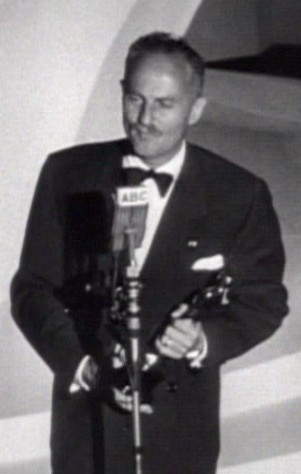 A man in a suit holding an award statuette stands at a podium delivering an acceptance speech.