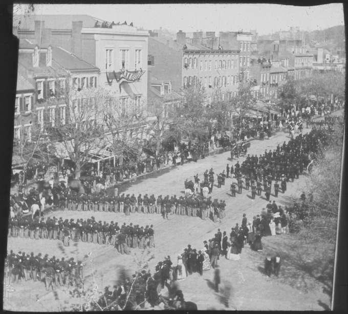 A procession of several hundred people moves down a street lined with tall buildings.