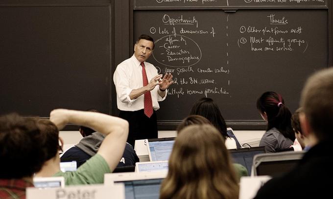 A professor stands in front of a classroom of students and delivers a lecture.