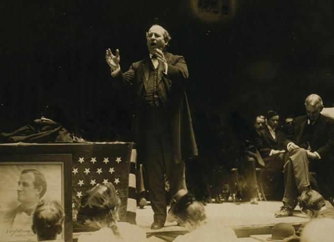 An old photograph of a man in a suit and tie speaking to an audience from a stage next to a display of the American flag.