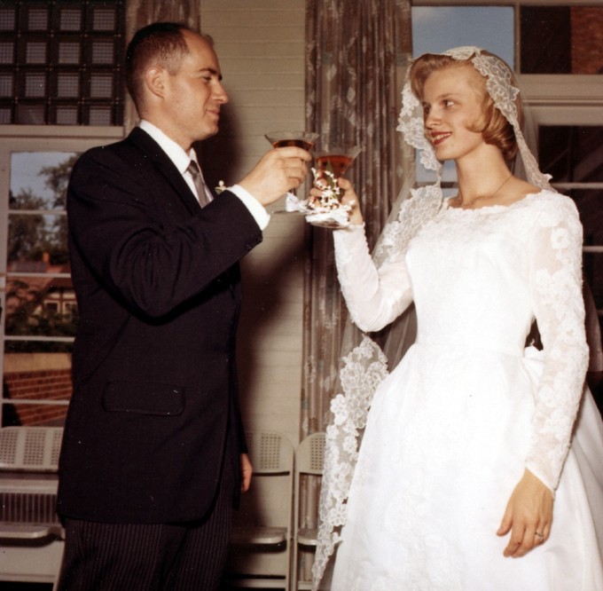 A man in a suit and a woman in a wedding dress each hold a glass and smile at one another while toasting.