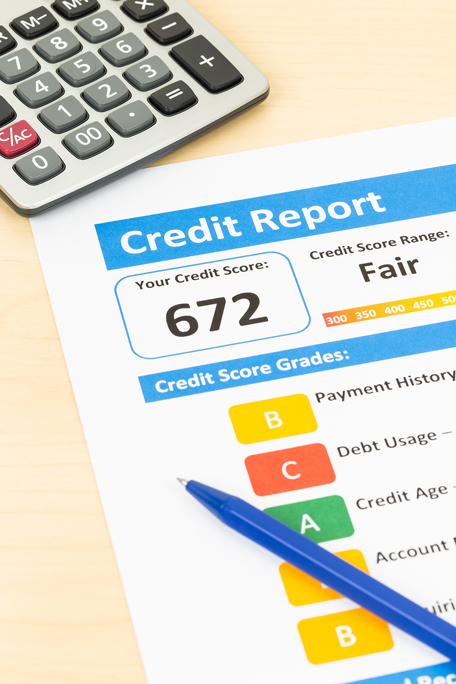 The image shows a printed credit report on a desk, partially covered by a calculator in the top-left corner and a blue pen resting on the paper. The report displays a credit score of 672, classified as ‘Fair’ on a scale from 300 to 850, with color-coded indicators for ranges such as Poor, Fair, Good, and Excellent. Below the score, a section titled ‘Credit Score Grades’ lists categories including Payment History, Debt Usage, Credit Age, and Account Mix, each with a color-coded grade ranging from A to C. The grades indicate the individual’s performance in different areas affecting the credit score.