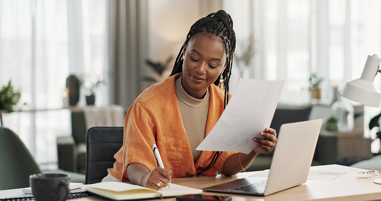The image shows a young woman sitting at a desk in a well-lit, modern workspace. She is holding a document in one hand while writing notes with a pen in the other. She is dressed in a casual yet professional outfit, wearing a turtleneck sweater and a loose-fitting cardigan. A laptop is open in front of her, along with a notebook, a coffee mug, and other office essentials. The background features large windows with natural light filtering through, along with houseplants and stylish decor, creating a warm and productive atmosphere. She has a focused and content expression, indicating engagement with her work.