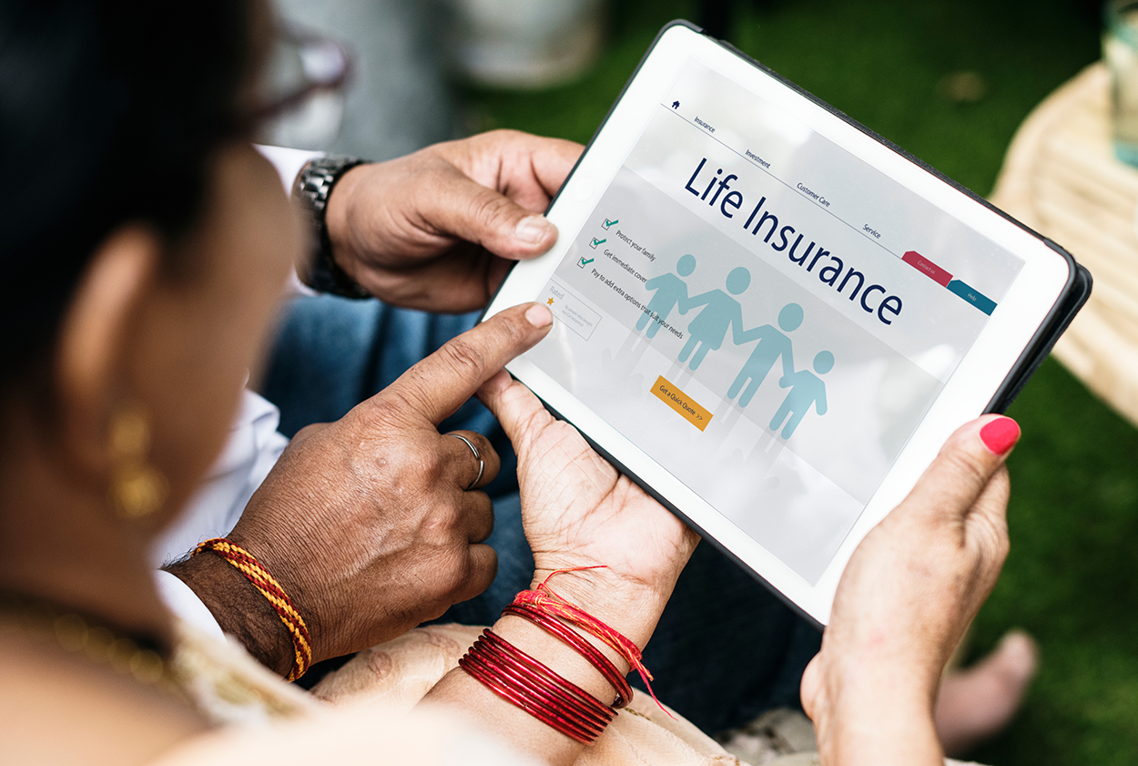 The image shows two people sitting outdoors and holding a tablet displaying a life insurance web page. The screen features the title “Life Insurance” along with an illustration of a family holding hands and a checklist highlighting benefits such as family protection and financial security. One person is pointing at the screen while the other holds the device, both appearing engaged in reviewing the information. They are wearing traditional bracelets and jewelry, suggesting a cultural or personal significance. The background is slightly blurred with a table and greenery visible, creating a relaxed and thoughtful setting.
