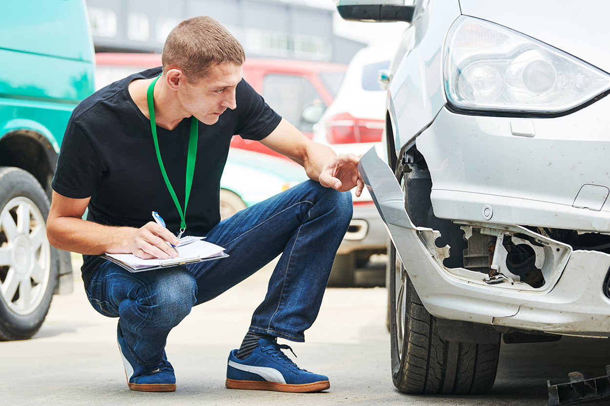 The image shows an insurance adjuster or claims inspector examining a damaged vehicle. He is crouching next to a car with a broken front bumper, taking notes on a clipboard, and inspecting the extent of the damage. He is wearing a lanyard, suggesting an official role. Several vehicles are visible in the background, indicating a repair shop or accident assessment site. The scene conveys the process of evaluating an auto insurance claim.