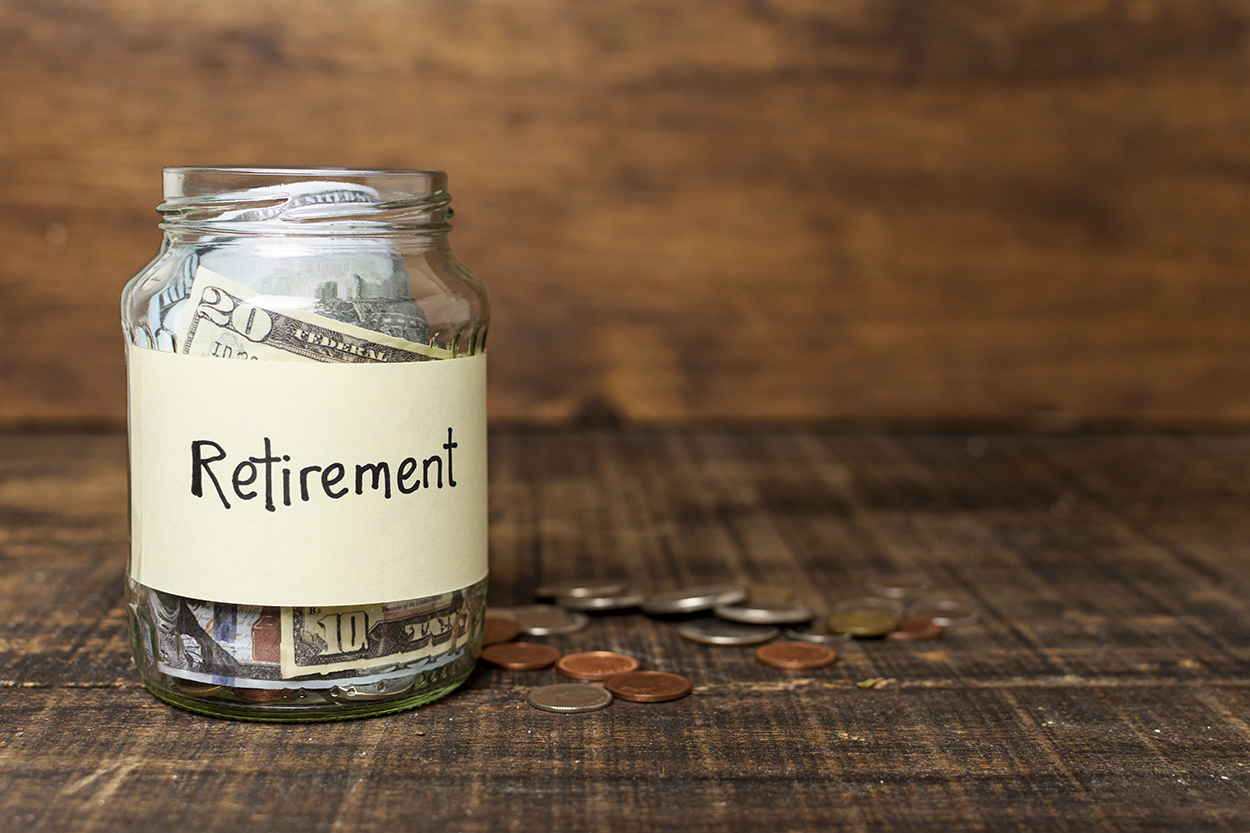 A glass jar labeled ‘Retirement’ filled with cash, with scattered coins on a wooden surface.
